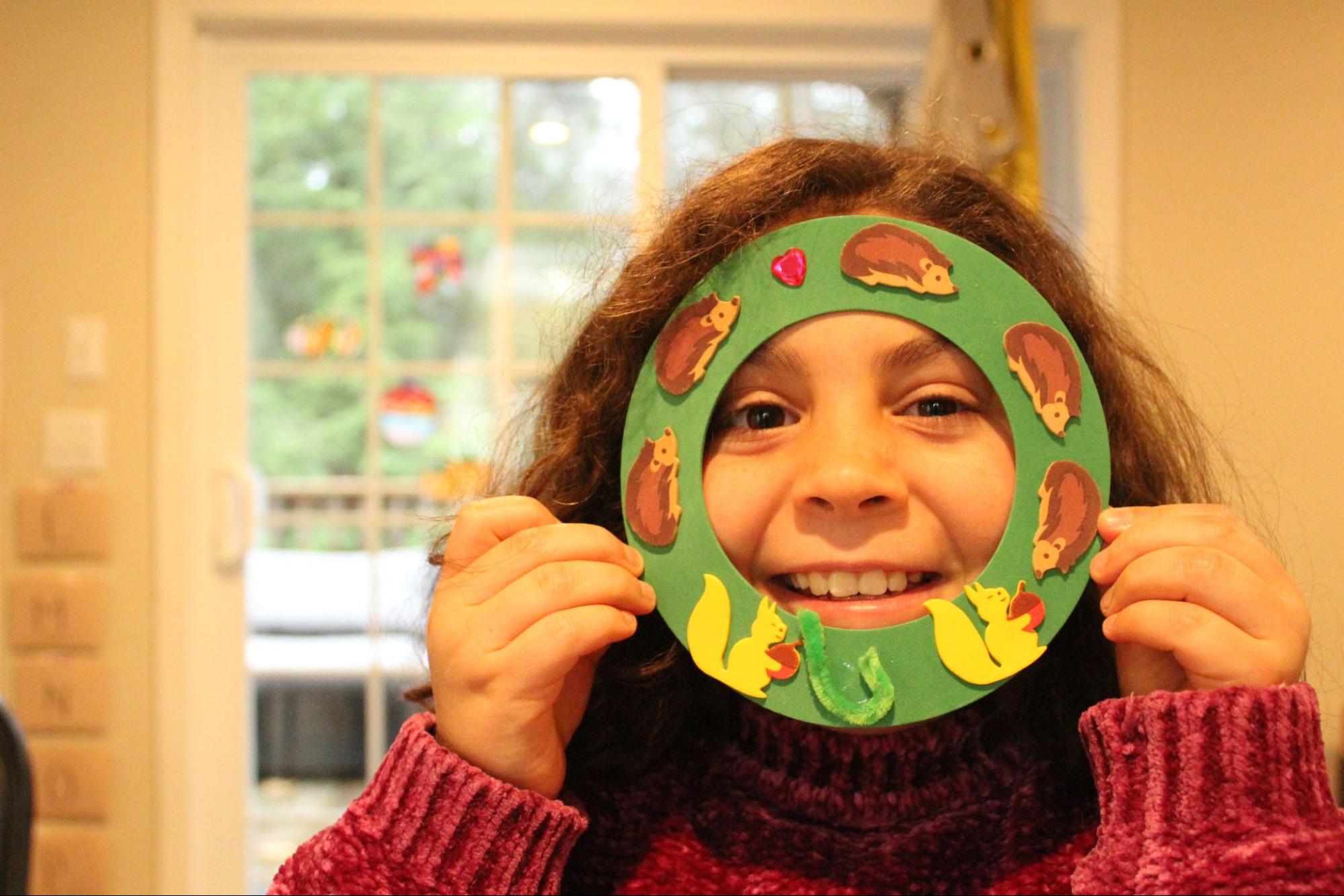 Young girl smiling through a circle craft she made with hedgehogs and squirrels on it